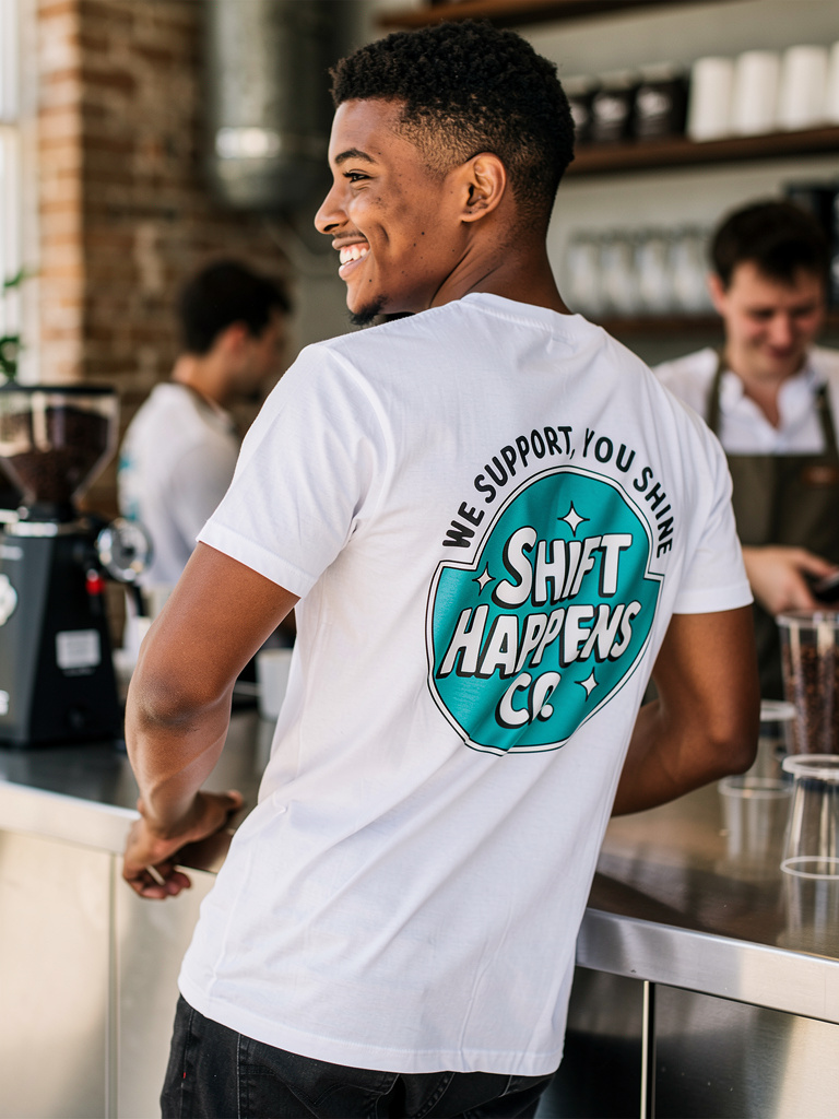 Person wearing a white t-shirt with a blue logo and text in a coffee shop setting